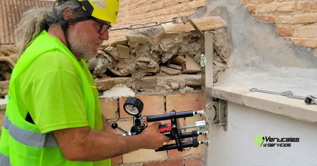 Primer plano de un operario realizando una prueba de carga o resistencia en un anclaje de seguridad instalado en una pared exterior. El técnico utiliza un extractor hidráulico con manómetro para verificar la fijación cerca de una zona de tejas y ladrillo.
