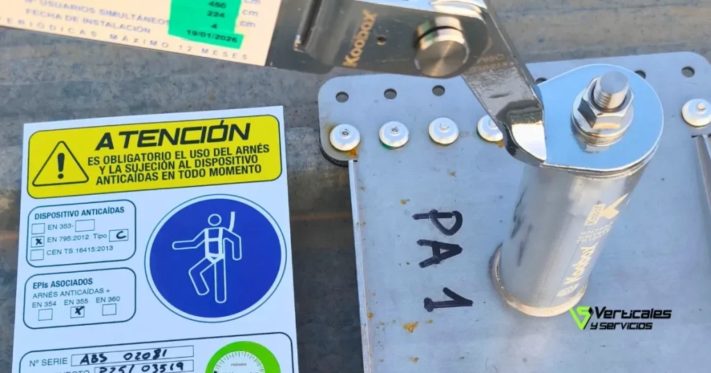 Detalle en primer plano de un poste de anclaje metálico instalado sobre una cubierta. Al lado se aprecia una etiqueta de señalización obligatoria que indica el cumplimiento de la normativa UNE-EN 795 Clase C, resaltando el uso obligatorio de arnés y sistemas anticaídas.